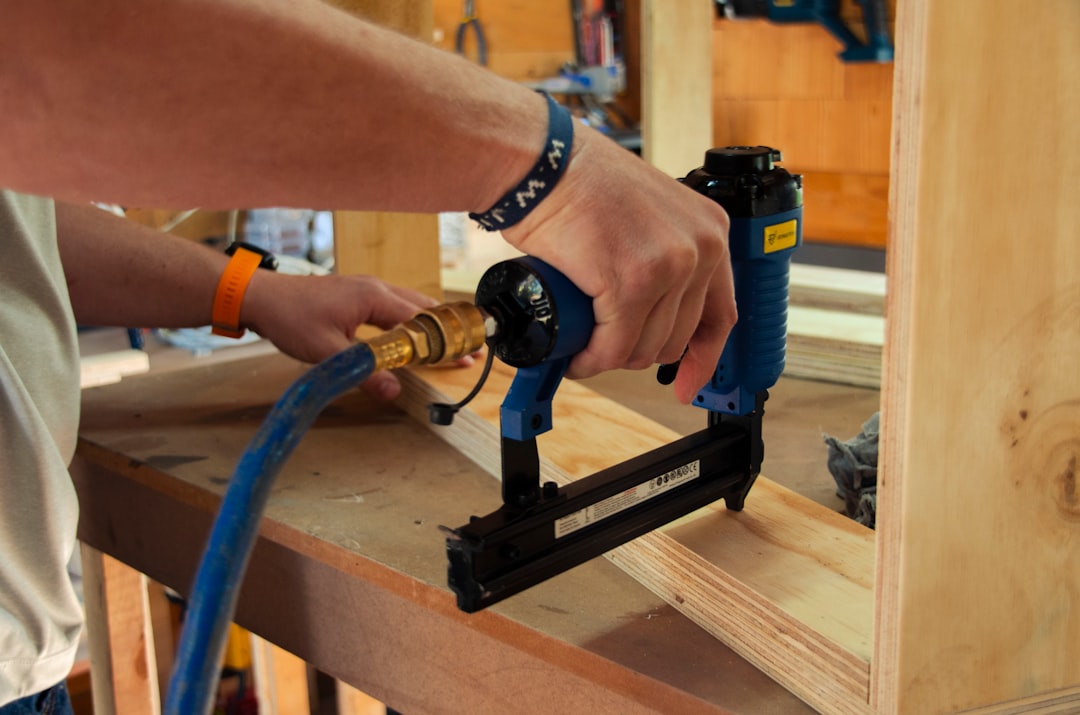 Close-up of a person using a blue pneumatic brad nailer to join plywood boards on a wooden workbench, with a blue air compressor hose attached. Man is wearing a blue bracelet with the chirsitan slogan letters W W J D in white.