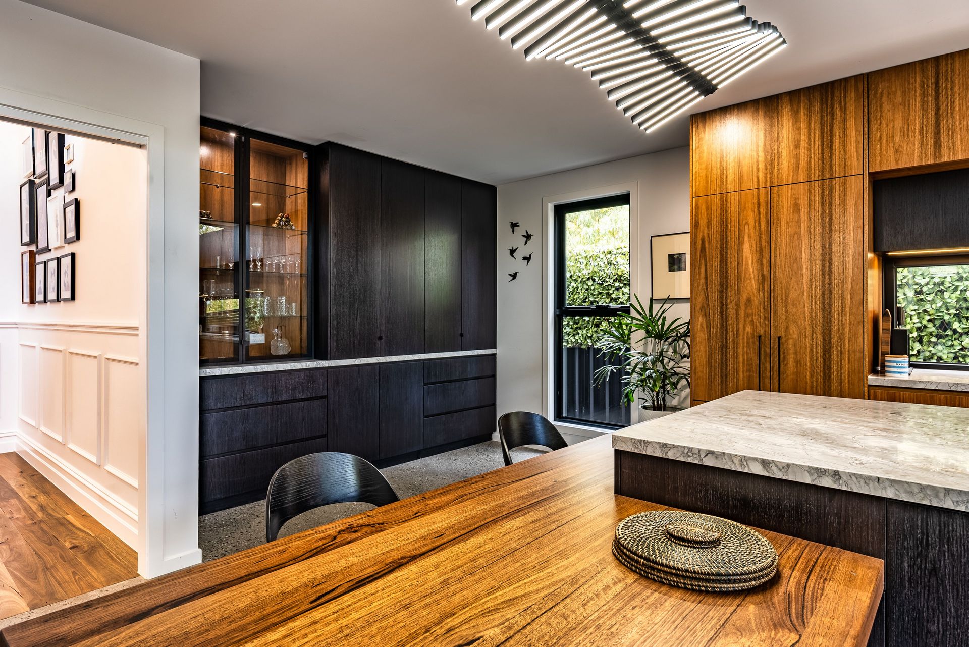 Contemporary open-plan kitchen and dining area featuring a large natural wood dining table, sleek black floor-to-ceiling cabinetry, and a marble-topped island. Large windows offer a view of green foliage, while a hallway with a gallery wall of framed pictures is visible in the background.